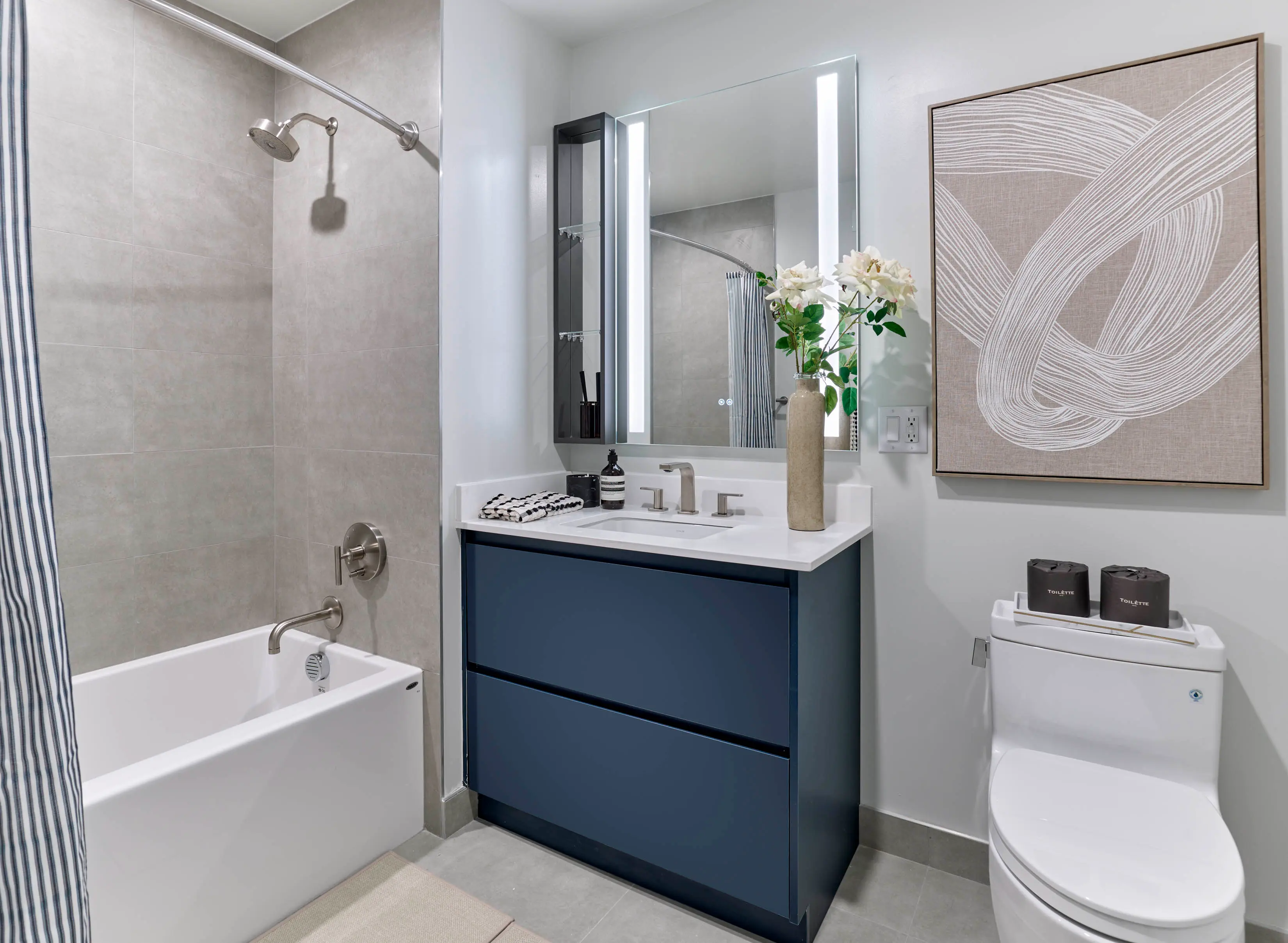 Modern apartment bathroom featuring a navy vanity, illuminated mirror, bathtub with showerhead, and neutral wall art